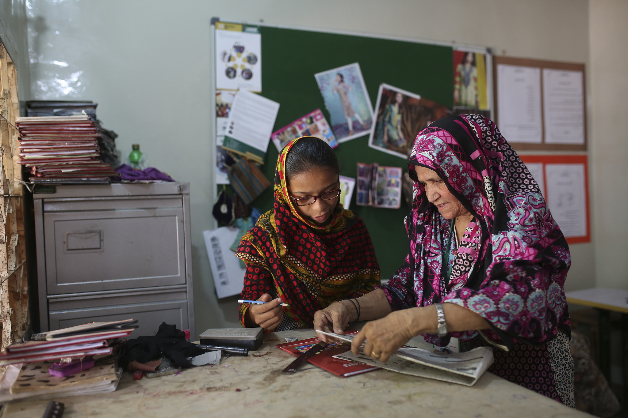 Woman teaching sewing design skills to a younger woman in front of pin board, Pakistan
